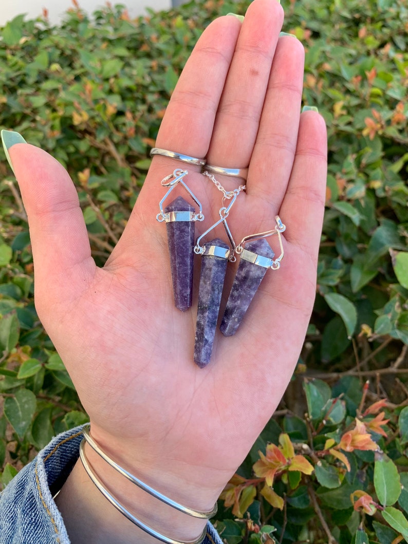 May include: Three purple amethyst crystal point pendants on silver chains. The pendants are hanging from a hand.