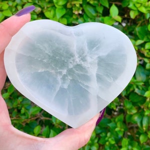 May include: A heart-shaped, white crystal dish with a translucent appearance. The dish has a smooth, polished surface and a natural, veined pattern. The background is blurred green foliage.