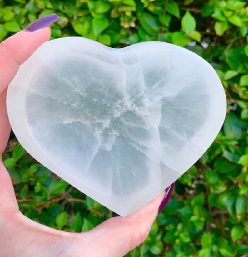 May include: A heart-shaped, white crystal dish with a translucent appearance. The dish has a smooth, polished surface and a natural, veined pattern. The background is blurred green foliage.