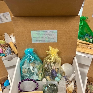 May include: An open box filled with a variety of crystals and stones, including selenite, rose quartz, and palo santo sticks. The box also includes a selenite ruler, a crystal bracelet, a palm stone, and a sphere with a stand. The box is labelled "Ultimate Crystal Trove (largest): 5-6 Selenite Ruler (Cleansing & recharging!) 1/2 lb of Random Tumbled Stones 1 lb Random Rough Stones 4 Palo Santo Sticks Rose Quartz Mini Point Mini Selenite Tower Palm Stone Magical Keychain Crystal Bracelet Handmade Pendant Sphere with Stand"