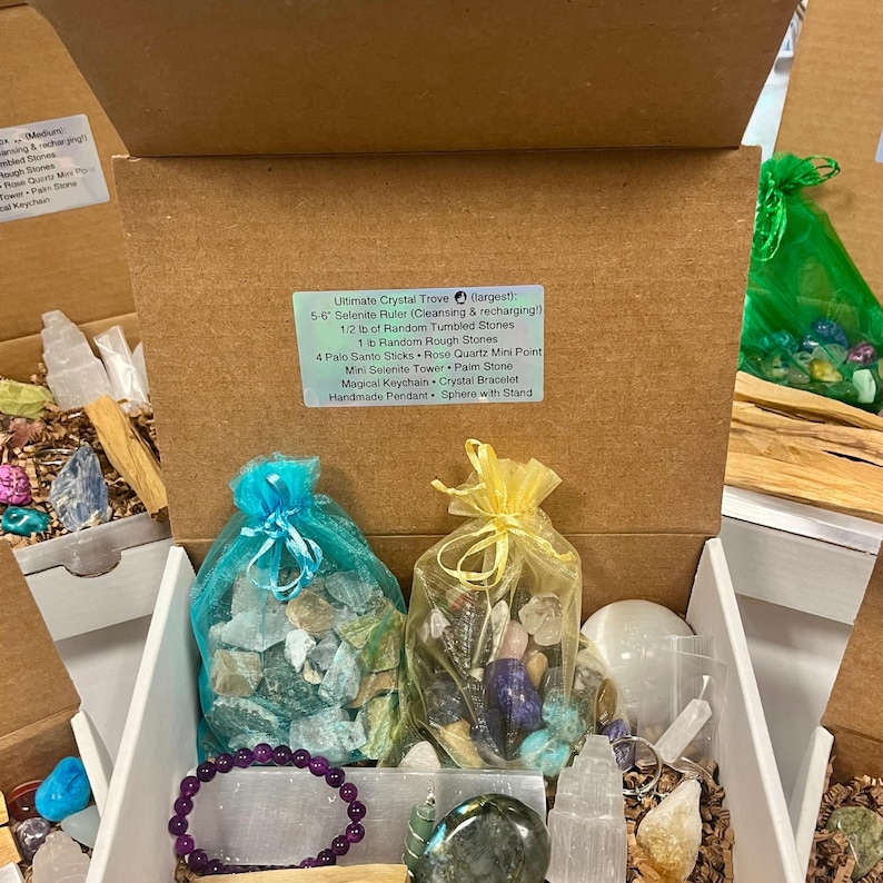 May include: An open box filled with a variety of crystals and stones, including selenite, rose quartz, and palo santo sticks. The box also includes a selenite ruler, a crystal bracelet, a palm stone, and a sphere with a stand. The box is labelled "Ultimate Crystal Trove (largest): 5-6 Selenite Ruler (Cleansing & recharging!) 1/2 lb of Random Tumbled Stones 1 lb Random Rough Stones 4 Palo Santo Sticks Rose Quartz Mini Point Mini Selenite Tower Palm Stone Magical Keychain Crystal Bracelet Handmade Pendant Sphere with Stand"