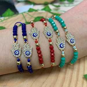 May include: Four beaded bracelets with a gold hamsa hand charm with a blue evil eye. The bracelets are blue, red, and green.