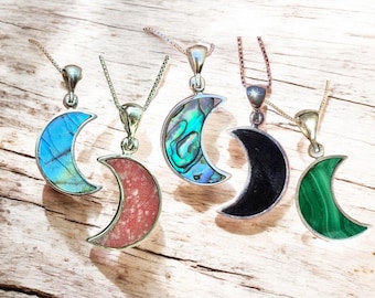 May include: Five crescent moon pendant necklaces with silver chains. The moon pendants feature various materials, including blue, red, abalone, black, and green. The necklaces are displayed on a light-colored wooden surface.