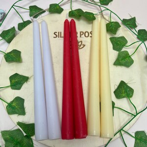 May include: Three taper candles in white, red, and cream colors. The candles are arranged on a wooden surface with green leaves.