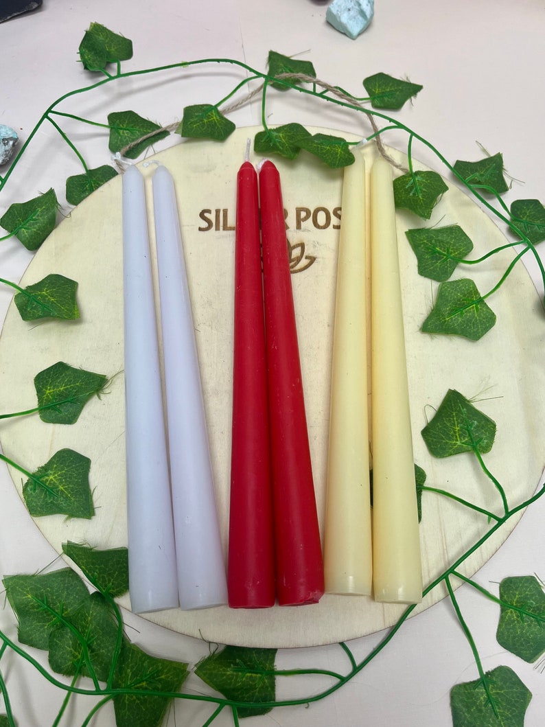 May include: Three taper candles in white, red, and cream colors. The candles are arranged on a wooden surface with green leaves.