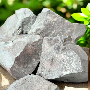 K&ouml;nnte beinhalten: Eine Ansammlung rauer, grauer H&auml;matitsteine mit metallischem Glanz. Die Steine haben gezackte Kanten und unterschiedliche Gr&ouml;&szlig;en mit einem nat&uuml;rlichen, erdigen Aussehen. Der Hintergrund zeigt verschwommenes gr&uuml;nes Laub.