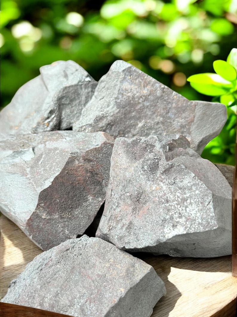 K&ouml;nnte beinhalten: Eine Ansammlung rauer, grauer H&auml;matitsteine mit metallischem Glanz. Die Steine haben gezackte Kanten und unterschiedliche Gr&ouml;&szlig;en mit einem nat&uuml;rlichen, erdigen Aussehen. Der Hintergrund zeigt verschwommenes gr&uuml;nes Laub.