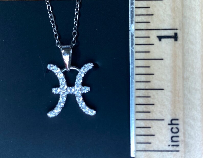 K&ouml;nnte beinhalten: Silberne Halskette mit einem Anh&auml;nger in Form des Sternzeichen Fische, besetzt mit klaren Zirkonia-Steinen. Der Anh&auml;nger ist etwa 2,5 cm lang.