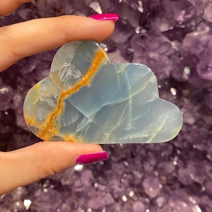 May include: A light blue cloud-shaped piece of polished blue calcite with gold veins. The stone is held in a hand with pink nail polish.