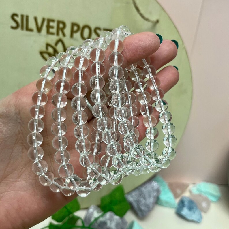 May include: Clear quartz crystal bead bracelets. The bracelets are made of round, transparent quartz beads strung together. The beads are held in a hand, with the text "SILVER POST" visible in the background.