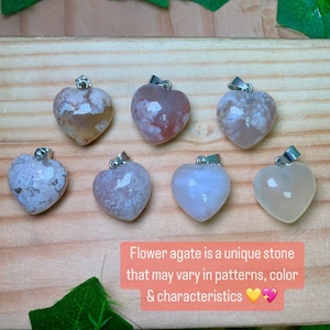 Puede incluir: Siete colgantes de &aacute;gata de flor en forma de coraz&oacute;n con engarces de plata. Las &aacute;gatas son todas de diferentes tonos de blanco, rosa y marr&oacute;n con patrones &uacute;nicos. El texto "El &aacute;gata de flor es una piedra &uacute;nica que puede variar en patrones, color y caracter&iacute;sticas" est&aacute; escrito debajo de los colgantes.