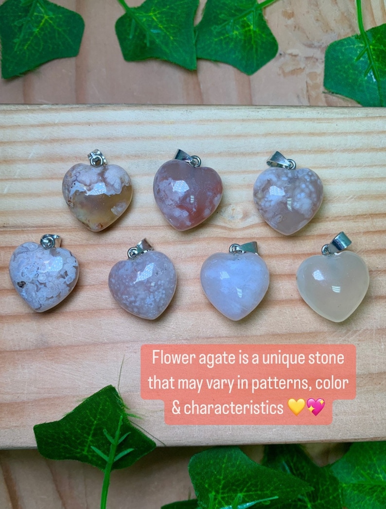 Puede incluir: Siete colgantes de &aacute;gata de flor en forma de coraz&oacute;n con engarces de plata. Las &aacute;gatas son todas de diferentes tonos de blanco, rosa y marr&oacute;n con patrones &uacute;nicos. El texto "El &aacute;gata de flor es una piedra &uacute;nica que puede variar en patrones, color y caracter&iacute;sticas" est&aacute; escrito debajo de los colgantes.