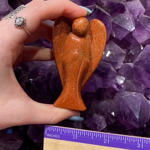May include: A small, brown, sandstone angel figurine. The angel is approximately 5 cm tall and is standing on a purple background.