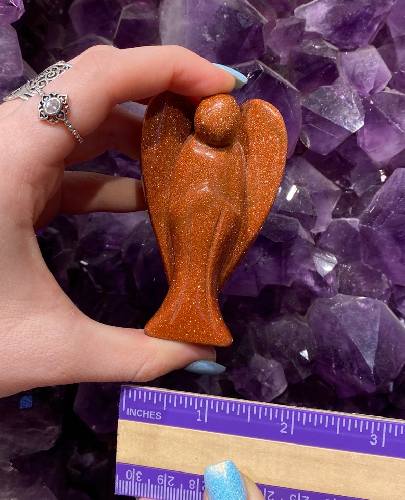May include: A small, brown, sandstone angel figurine. The angel is approximately 5 cm tall and is standing on a purple background.