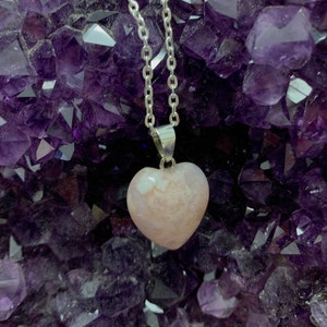 Puede incluir: Un collar de cadena de plata con un colgante en forma de coraz&oacute;n rosa. El colgante est&aacute; hecho de una piedra transl&uacute;cida con remolinos blancos. El collar se muestra sobre un lecho de cristales de amatista p&uacute;rpura.