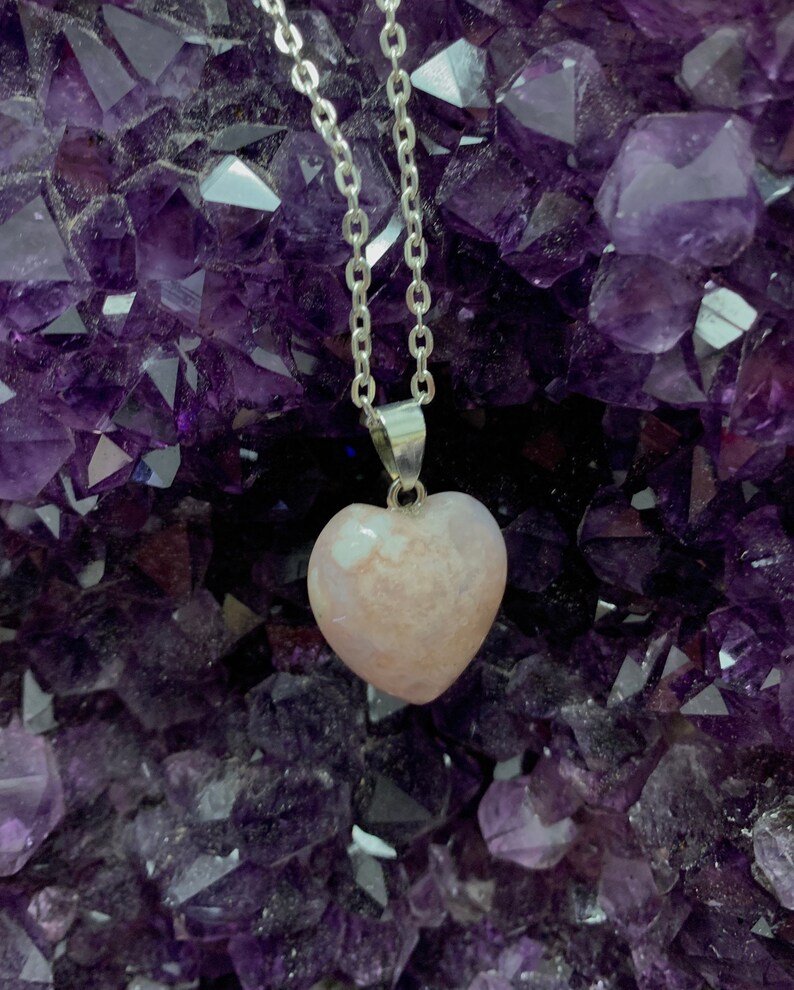 Puede incluir: Un collar de cadena de plata con un colgante en forma de coraz&oacute;n rosa. El colgante est&aacute; hecho de una piedra transl&uacute;cida con remolinos blancos. El collar se muestra sobre un lecho de cristales de amatista p&uacute;rpura.
