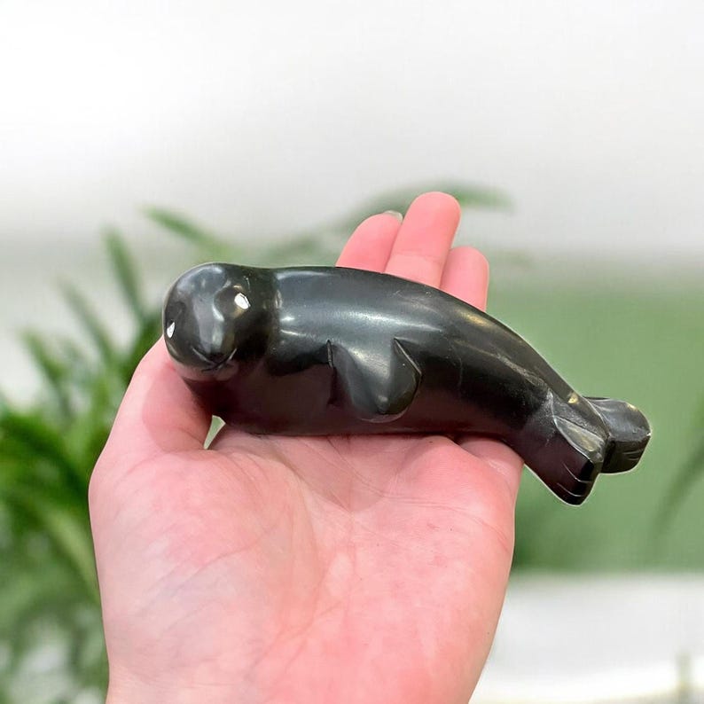 May include: A black, hand-carved soapstone sculpture of a seal. The seal is lying on its belly with its head turned to the side. The sculpture is detailed with small white dots for eyes.