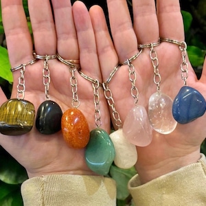 May include: A collection of gemstone keychains in various colors, including yellow, black, orange, green, white, pink, clear, and blue. Each stone is attached to a silver chain and keyring. The keychains are held in a person's hands.