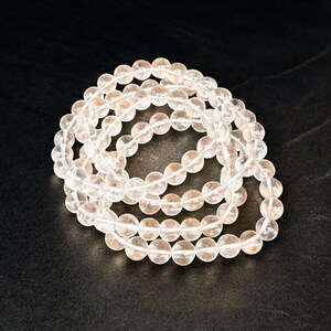 May include: A collection of clear, round crystal beads strung together, forming multiple strands. The beads are translucent and appear to be made of quartz. The arrangement is on a dark, textured surface, creating a contrast with the light-colored beads.