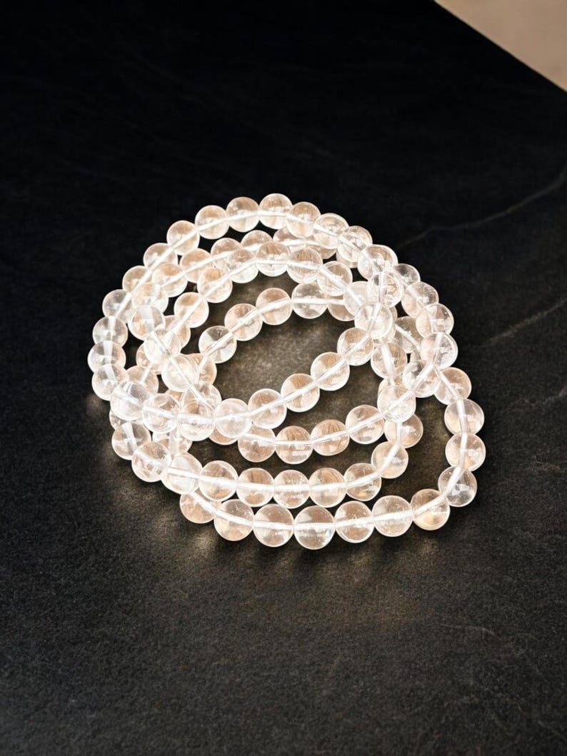 May include: A collection of clear, round crystal beads strung together, forming multiple strands. The beads are translucent and appear to be made of quartz. The arrangement is on a dark, textured surface, creating a contrast with the light-colored beads.