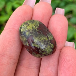 May include: A smooth, polished green and red gemstone with a mottled pattern. The stone is oval-shaped and has a glossy finish.