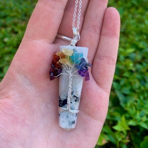May include: A silver chain necklace with a white crystal pendant featuring a wire-wrapped tree of life design with colorful gemstones. The gemstones are arranged in a rainbow pattern, representing the seven chakras.