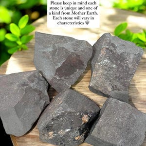 K&ouml;nnte beinhalten: Nahaufnahme von mehreren dunkelgrauen Steinen mit rauer Textur und unterschiedlichen Formen. Einige Steine weisen sichtbare Mineralablagerungen auf. Der Text im Bild lautet: "Bitte beachten Sie, dass jeder Stein einzigartig ist und von Mutter Erde stammt."