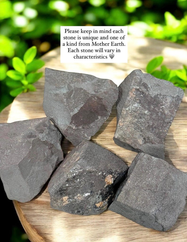 K&ouml;nnte beinhalten: Nahaufnahme von mehreren dunkelgrauen Steinen mit rauer Textur und unterschiedlichen Formen. Einige Steine weisen sichtbare Mineralablagerungen auf. Der Text im Bild lautet: "Bitte beachten Sie, dass jeder Stein einzigartig ist und von Mutter Erde stammt."
