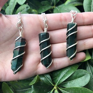 May include: Three green and red gemstone pendants wrapped in silver wire on silver chains. The pendants are all the same shape and size, and they are hanging from a hand.