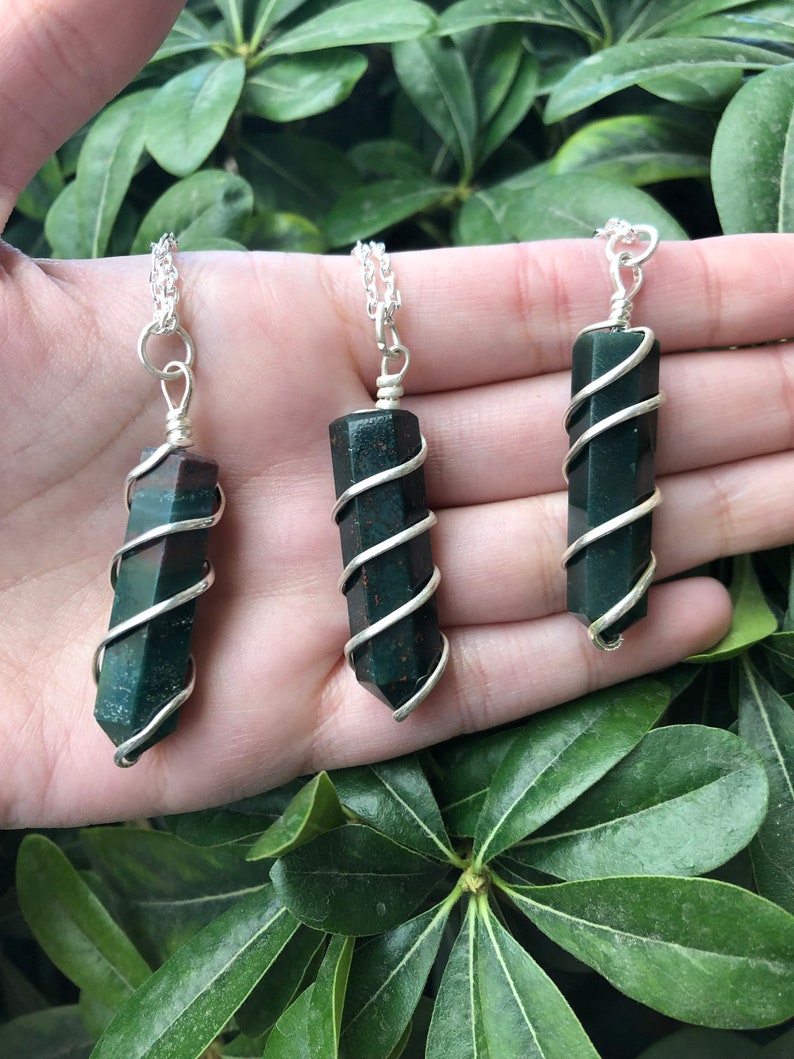 May include: Three green and red gemstone pendants wrapped in silver wire on silver chains. The pendants are all the same shape and size, and they are hanging from a hand.