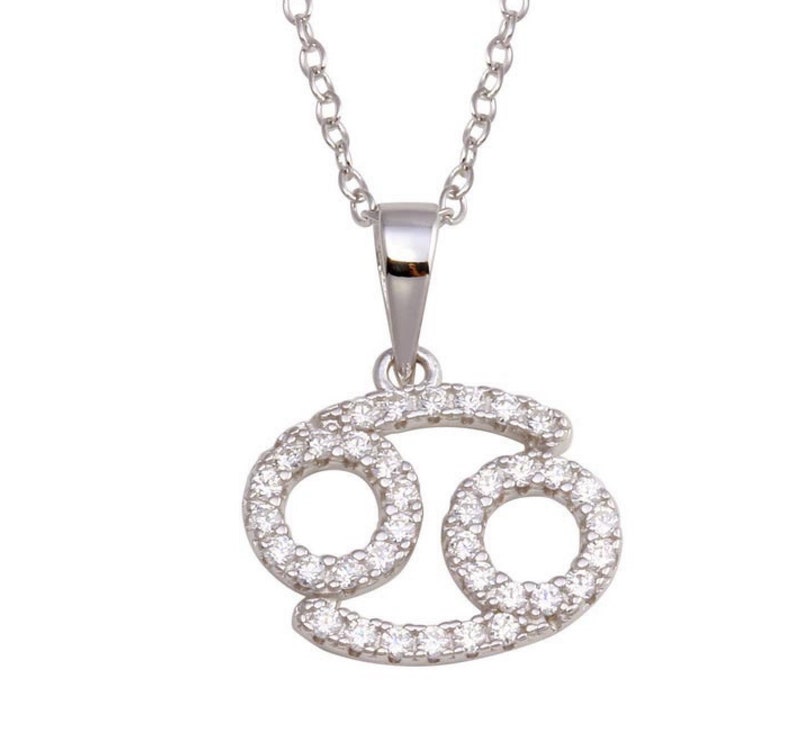 May include: Silver necklace with a pendant shaped like the Cancer zodiac sign. The pendant is encrusted with small, clear cubic zirconia stones.