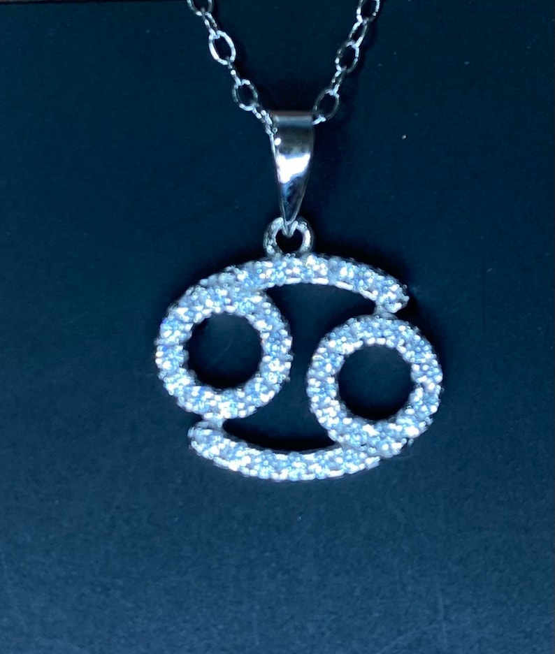 May include: Silver pendant necklace featuring a Cancer zodiac sign with a sparkling crystal design.