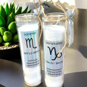 Puede incluir: Dos tarros de velas de vidrio con velas blancas y signos del zod&iacute;aco en las etiquetas. El tarro de la izquierda tiene un signo de Escorpio con las fechas del 23 de octubre al 21 de noviembre y el texto "INTENSO APASIONADO MISTERIOSO". El tarro de la derecha tiene un signo de Capricornio con las fechas del 22 de diciembre al 19 de enero y el texto "RESPONSABLE DISCIPLINADO AMBICIOSO".
