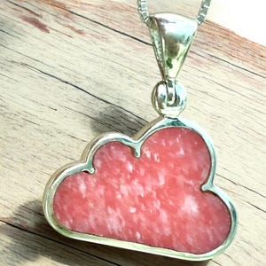 May include: A cloud-shaped pendant with a silver-colored frame and bail. The pendant's center is a pink stone with white marbling. The necklace chain is also silver-colored. The pendant is displayed on a wooden surface.