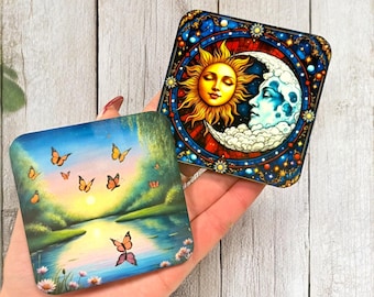 May include: Two square coasters with colorful designs. One features a sunset scene with butterflies, the other a sun and moon design. The coasters are held in a hand against a light wood background.