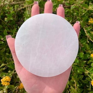 May include: A circular, white selenite charging plate. The plate has a smooth surface with natural veining. The plate is held in a hand, with a backdrop of green foliage and yellow flowers.