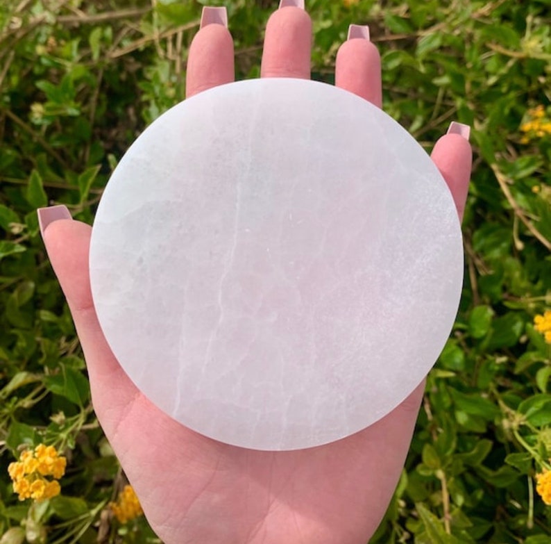 May include: A circular, white selenite charging plate. The plate has a smooth surface with natural veining. The plate is held in a hand, with a backdrop of green foliage and yellow flowers.