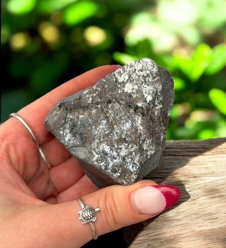 K&ouml;nnte beinhalten: Ein dunkelgrauer, unregelm&auml;&szlig;ig geformter Stein mit silbernen Flecken wird in einer Hand gehalten. Der Stein hat eine raue Textur und scheint ein Mineralexemplar zu sein. Ein silberner Ring mit einem Schildkr&ouml;ten-Design befindet sich am Finger der Person.