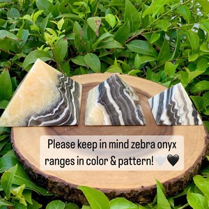 May include: Three zebra onyx pyramids with black, white, and tan patterns are displayed on a wooden slice. The pyramids are set against green foliage. Text on the image reads: "Please keep in mind zebra onyx ranges in colour & pattern!"