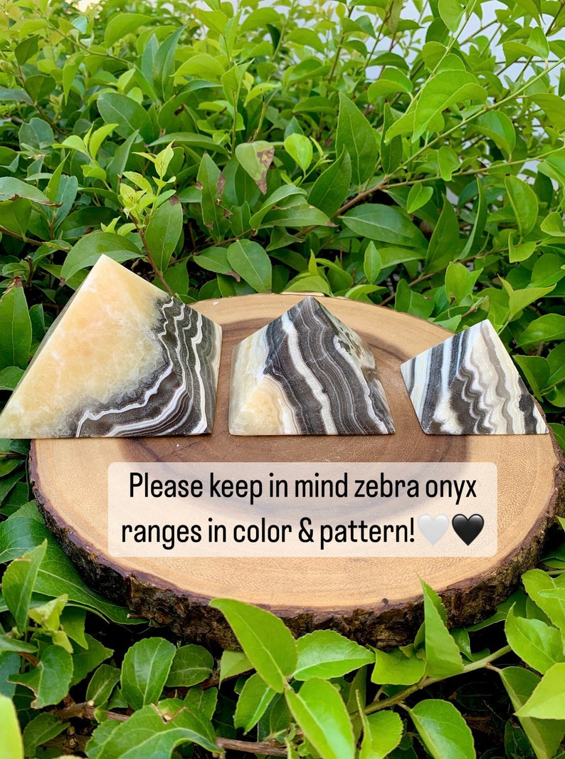 May include: Three zebra onyx pyramids with black, white, and tan patterns are displayed on a wooden slice. The pyramids are set against green foliage. Text on the image reads: "Please keep in mind zebra onyx ranges in colour & pattern!"