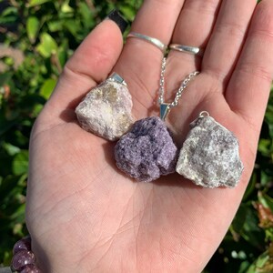 Op de afbeelding: Drie ruwe, natuurlijke lepidolietkristal hangers aan een zilveren ketting. De kristallen zijn in verschillende tinten paars, wit en grijs.
