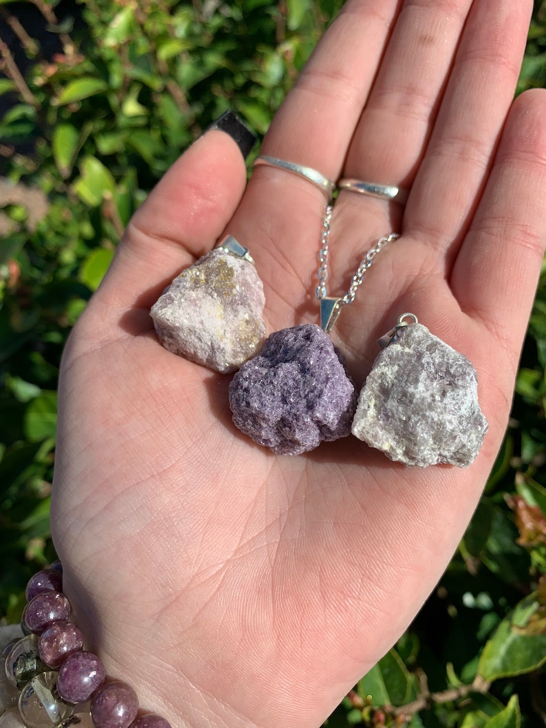 Op de afbeelding: Drie ruwe, natuurlijke lepidolietkristal hangers aan een zilveren ketting. De kristallen zijn in verschillende tinten paars, wit en grijs.
