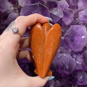 May include: A polished, orange-brown, heart-shaped angel figurine made of sunstone. The figurine has a sparkly, glittery texture.