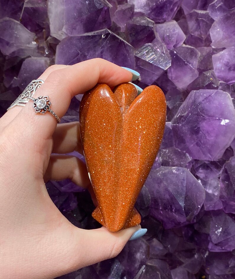 May include: A polished, orange-brown, heart-shaped angel figurine made of sunstone. The figurine has a sparkly, glittery texture.