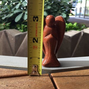May include: A red-brown sandstone angel figurine, approximately 6.5 cm tall, standing on a wooden surface. A yellow measuring tape is visible to the left of the figurine.