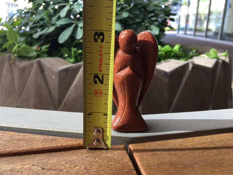 May include: A red-brown sandstone angel figurine, approximately 6.5 cm tall, standing on a wooden surface. A yellow measuring tape is visible to the left of the figurine.