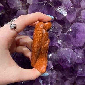 May include: A carved sandstone sculpture of a person with their arms crossed, standing on a bed of amethyst crystals. The sculpture is a reddish-brown colour.