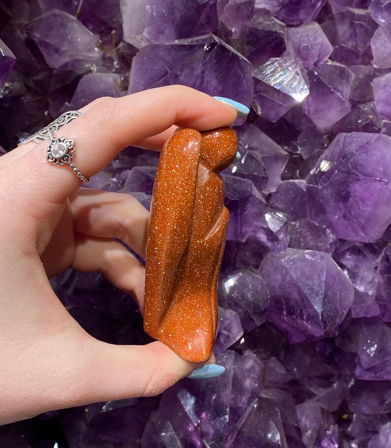 May include: A carved sandstone sculpture of a person with their arms crossed, standing on a bed of amethyst crystals. The sculpture is a reddish-brown colour.