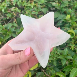 May include: A star-shaped crystal sculpture in a pale pink hue. The five points of the star are clearly defined, with a smooth, polished finish. The crystal is held against a backdrop of green leaves, appearing translucent with color variations.