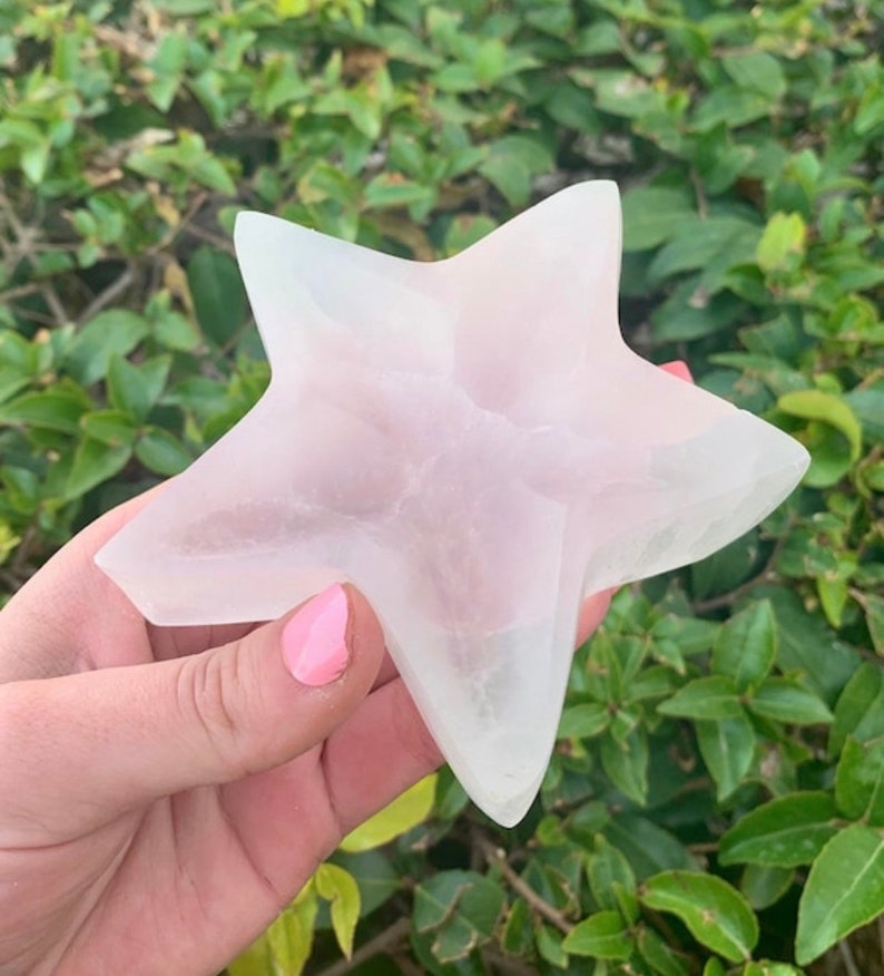 May include: A star-shaped crystal sculpture in a pale pink hue. The five points of the star are clearly defined, with a smooth, polished finish. The crystal is held against a backdrop of green leaves, appearing translucent with color variations.